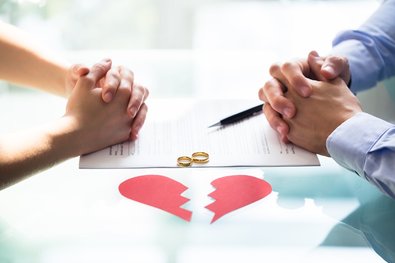 Couple's Hand On Divorce Paper With Wedding Rings And Broken Heart